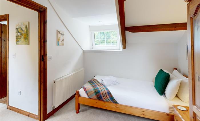 A bright twin bedroom with sloping ceiling, wooden furniture, tartan throw, and a window overlooking greenery.